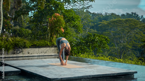 European brunette sports girl makes yoga in nature with a green rainforest on a lit background by the pool on a mat and breathes deeply.