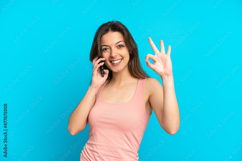 Young caucasian woman using mobile phone isolated on blue background showing ok sign with fingers Young caucasian woman using mobile phone isolated on blue background showing ok sign with fingers