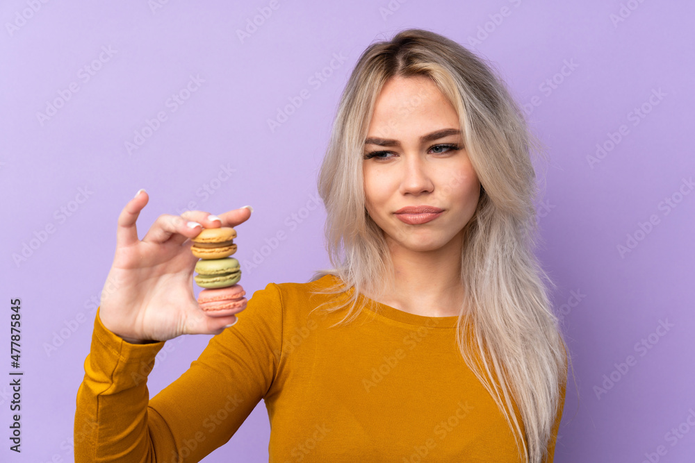 Teenager girl over isolated purple background holding colorful French macarons with sad expression Teenager girl over isolated purple background holding colorful French macarons with sad expression