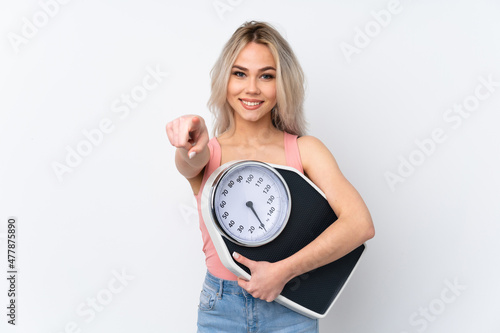 Teenager girl over isolated white background holding a weighing machine and pointing to the front