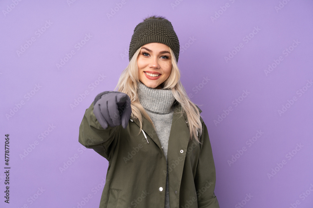 Teenager blonde girl with winter hat over isolated purple background points finger at you with a confident expression Teenager blonde girl with winter hat over isolated purple background points finger at you with a confident expression