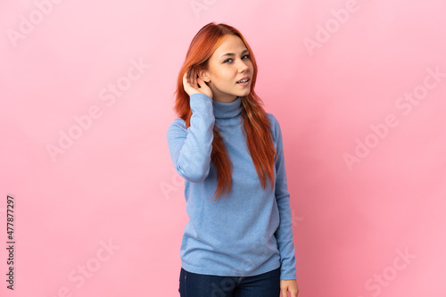Teenager Russian girl isolated on pink background listening to something by putting hand on the ear © luismolinero