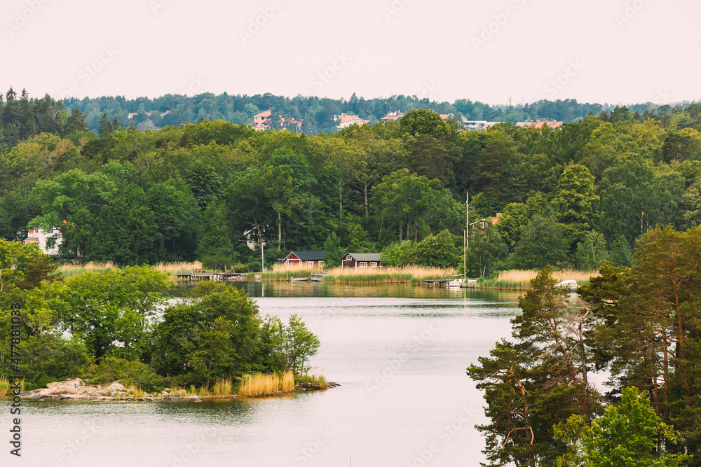 Sweden. Beautiful Swedish Wooden Log Cabins Houses On Coast In Summer Day. Lake Or River Landscape