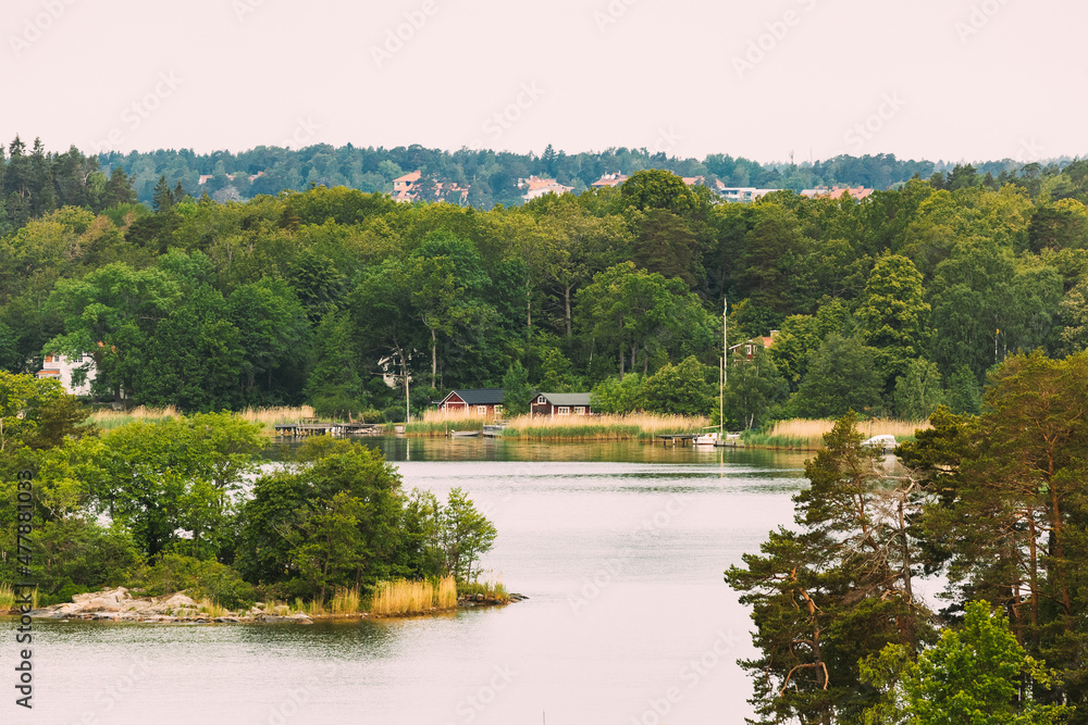 Sweden. Beautiful Swedish Wooden Log Cabins Houses On Coast In Summer Day. Lake Or River Landscape Sweden. Beautiful Swedish Wooden Log Cabins Houses On Coast In Summer Day. Lake Or River Landscape