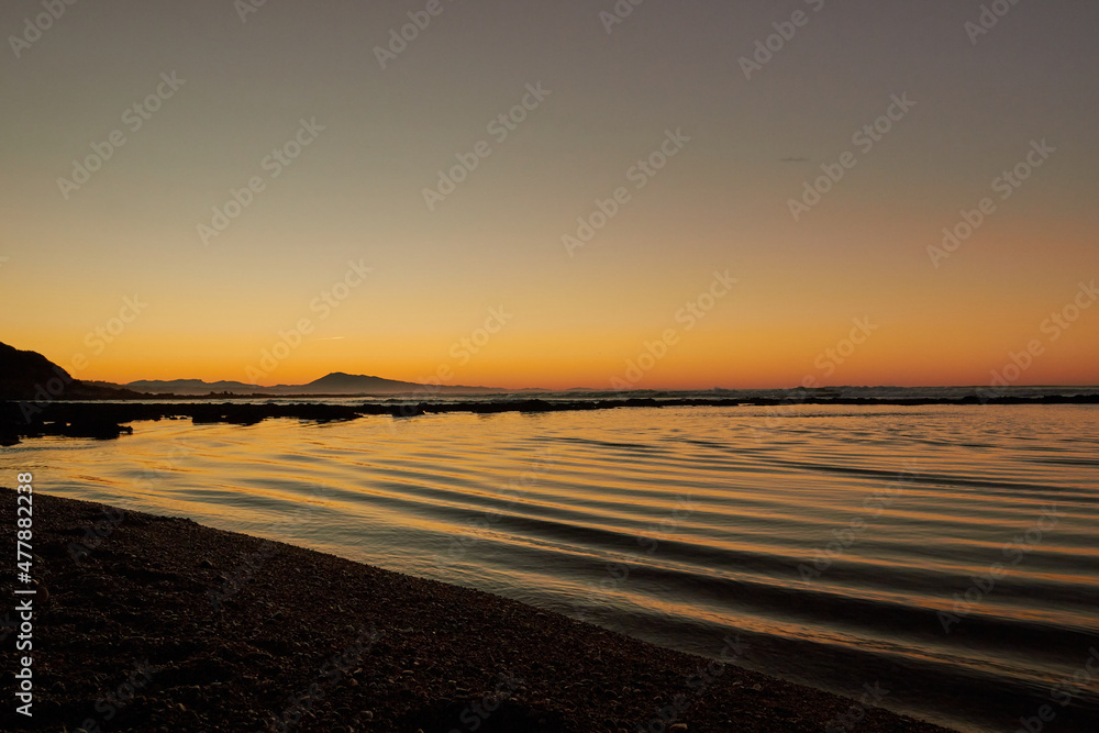 landscape of a beach with the waves hitting the rocks while the sun sets behind the mountains in the background, the orange light and the sound of the waves create a super relaxing scene, people walki landscape of a beach with the waves hitting the rocks while the sun sets behind the mountains in the background, the orange light and the sound of the waves create a super relaxing scene, people walki