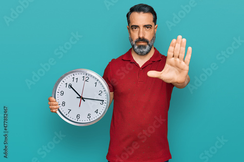 Middle aged man with beard holding big clock with open hand doing stop sign with serious and confident expression, defense gesture