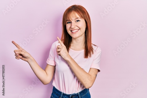 Redhead young woman wearing casual pink t shirt smiling and looking at the camera pointing with two hands and fingers to the side.