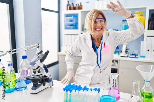 Middle age blonde woman working at scientist laboratory very happy and smiling looking far away with hand over head. searching concept.