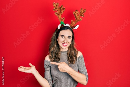 Young hispanic girl wearing deer christmas hat amazed and smiling to the camera while presenting with hand and pointing with finger.