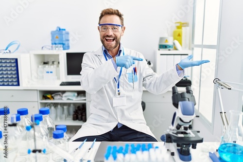 Middle age man working at scientist laboratory amazed and smiling to the camera while presenting with hand and pointing with finger.