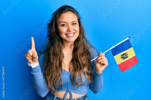 Young hispanic girl holding moldova flag smiling with an idea or question pointing finger with happy face, number one
