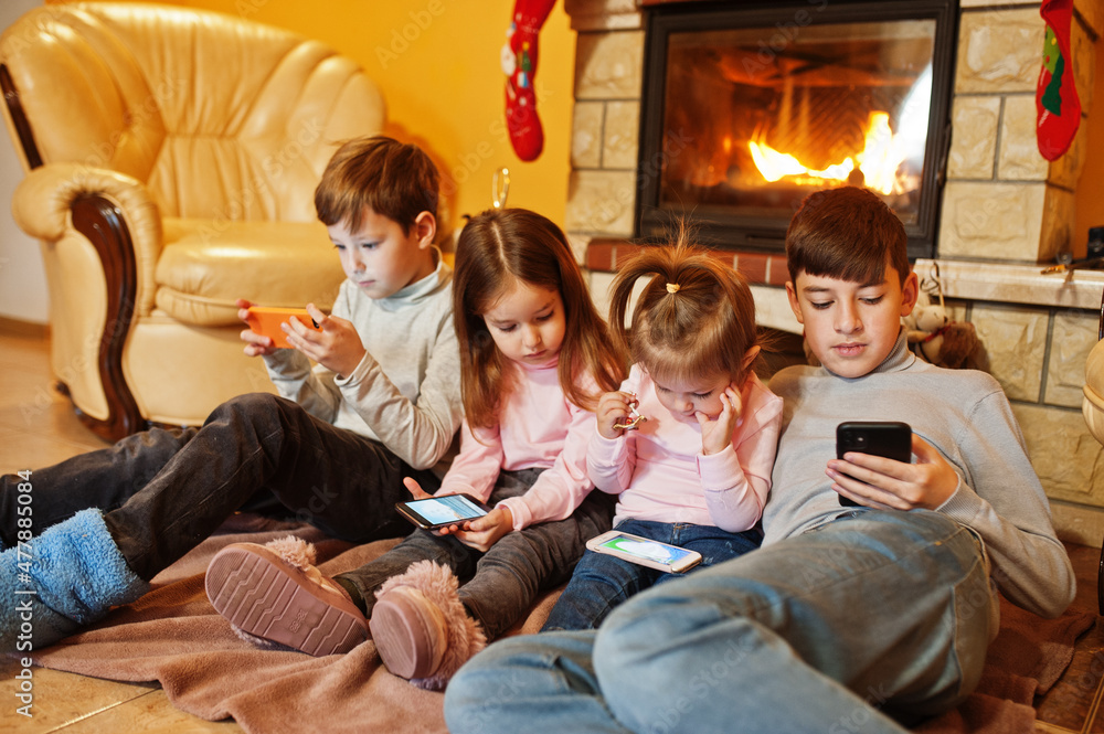 Happy four kids watching in they mobile phone at home by a fireplace in warm living room on winter day.