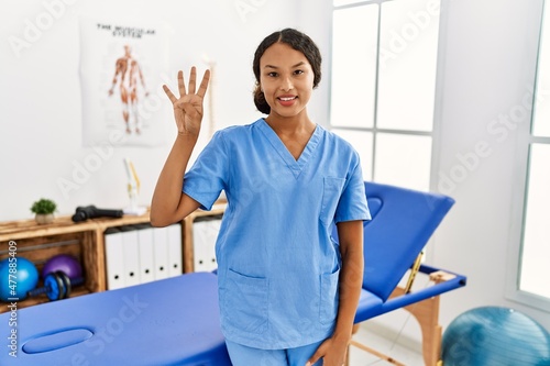 Beautiful hispanic physiotherapist woman at pain recovery clinic showing and pointing up with fingers number four while smiling confident and happy.