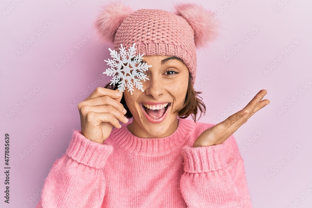 Young brunette woman holding snowflake wearing winter cap celebrating achievement with happy smile and winner expression with raised hand