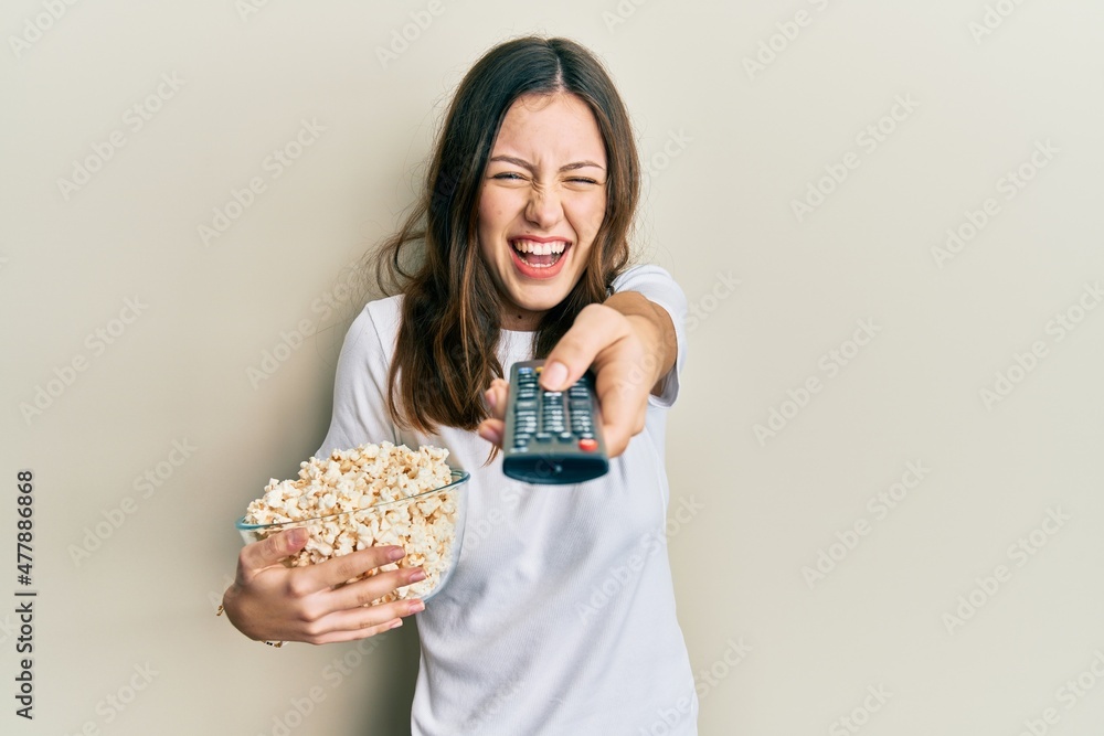 Young brunette woman eating popcorn using tv control smiling and laughing hard out loud because funny crazy joke.