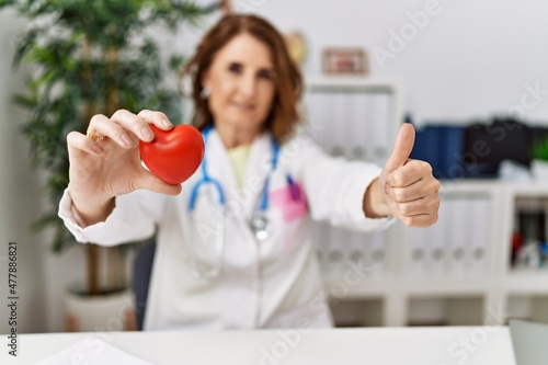 Middle age doctor woman holding heart at the clinic feeling unwell and coughing as symptom for cold or bronchitis. health care concept.