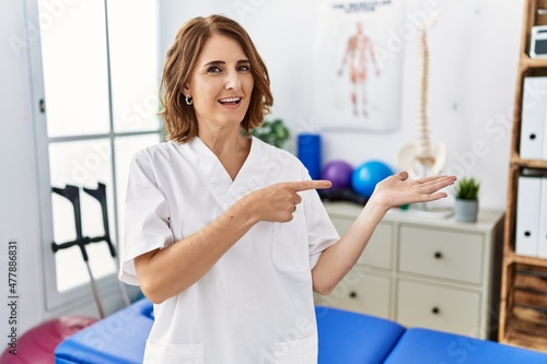 Middle age physiotherapist woman working at pain recovery clinic amazed and smiling to the camera while presenting with hand and pointing with finger.