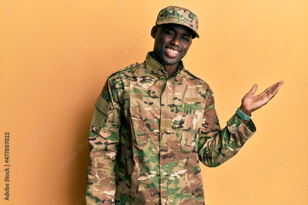 Young african american man wearing army uniform smiling cheerful presenting and pointing with palm of hand looking at the camera. Young african american man wearing army uniform smiling cheerful presenting and pointing with palm of hand looking at the camera.