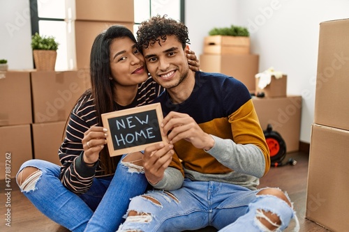 Young latin couple holding new home blackboard sitting on the floor.