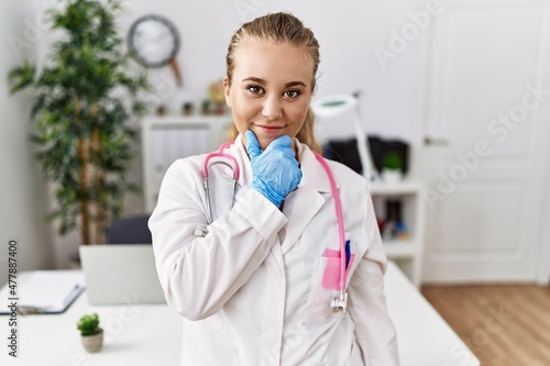 Young caucasian woman wearing doctor uniform and stethoscope at the clinic looking confident at the camera with smile with crossed arms and hand raised on chin. thinking positive.