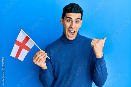 Handsome hispanic man holding england flag pointing thumb up to the side smiling happy with open mouth