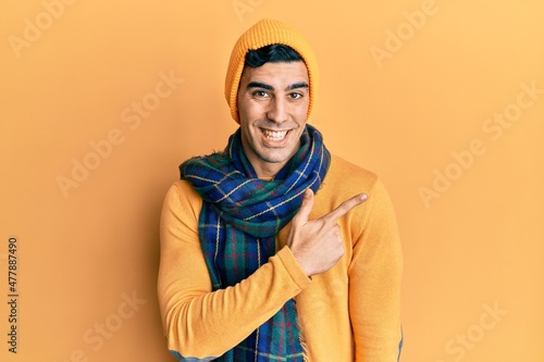Handsome hispanic man wearing wool hat and winter scarf cheerful with a smile of face pointing with hand and finger up to the side with happy and natural expression on face © Krakenimages.com