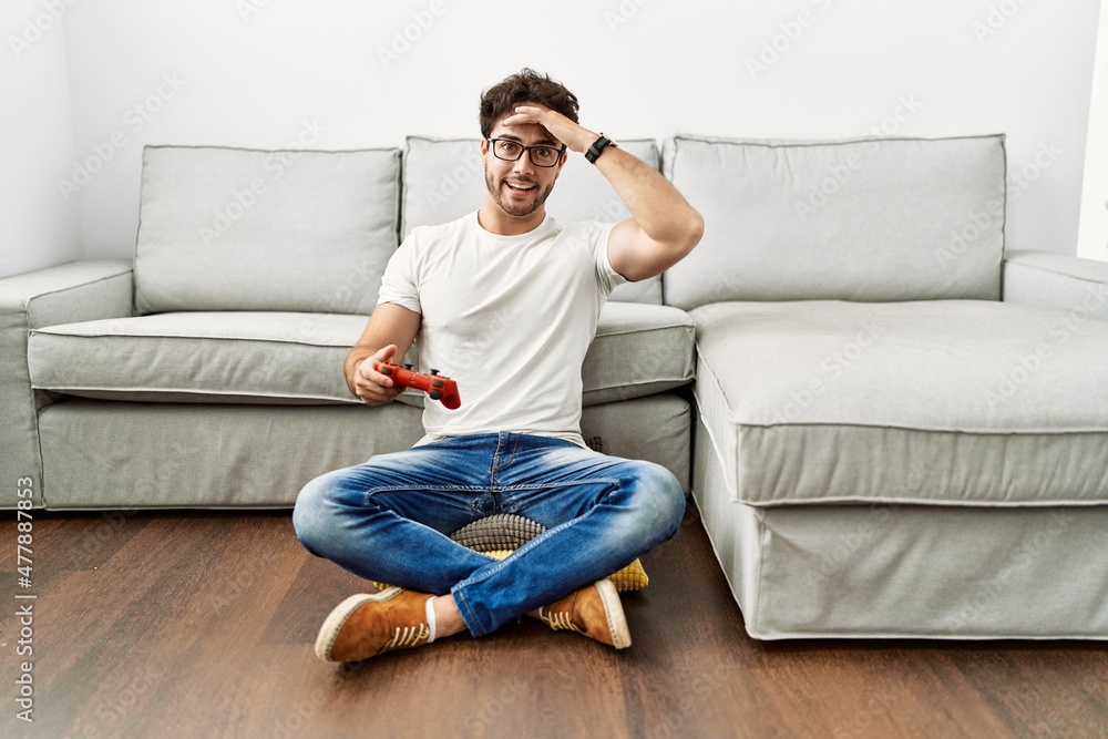 Hispanic man playing video game holding controller by the sofa stressed and frustrated with hand on head, surprised and angry face
