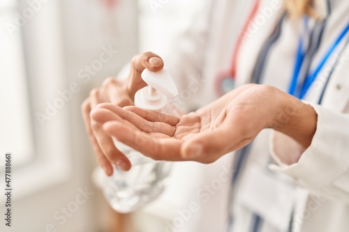 Young blonde woman wearing doctor uniform using sanitizer gel hands at clinic