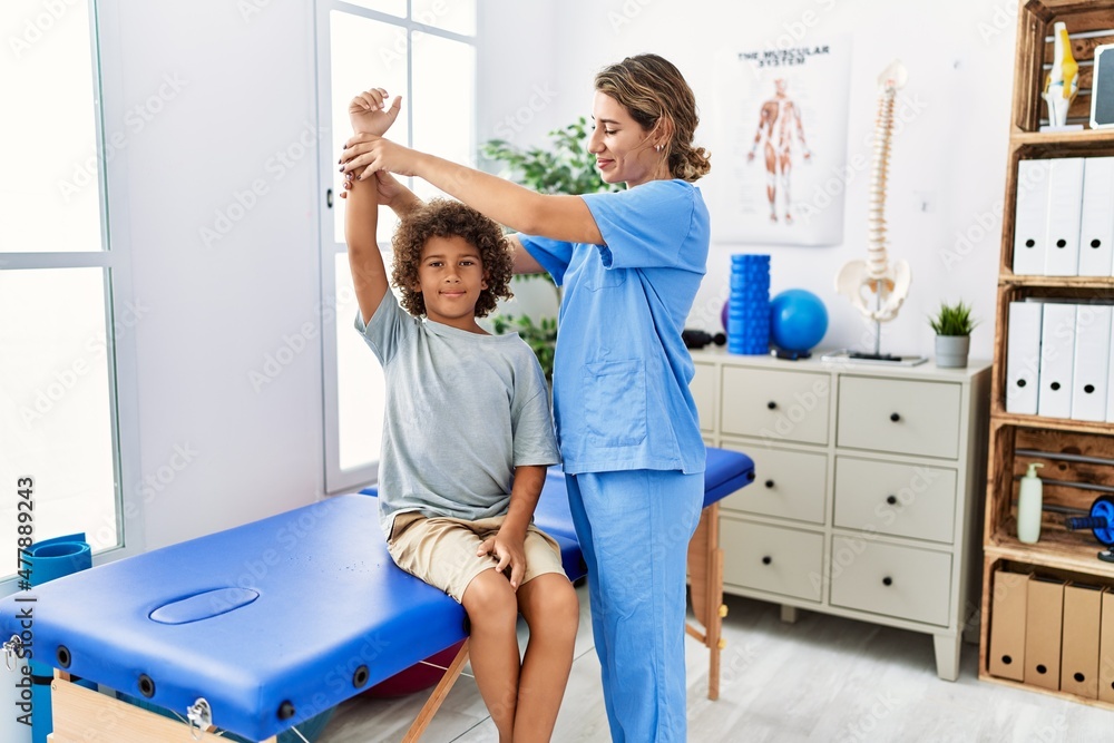 Obraz premium Mother and son wearing physiotherapist uniform having rehab session stretching arm at physiotherapy clinic