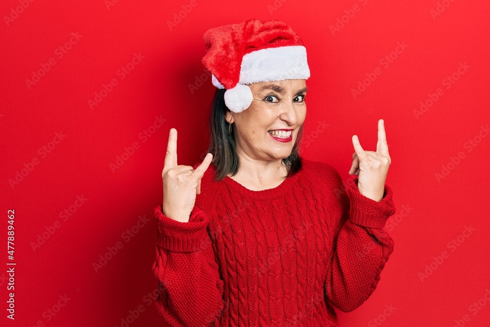Middle age hispanic woman wearing christmas hat shouting with crazy expression doing rock symbol with hands up. music star. heavy concept. Middle age hispanic woman wearing christmas hat shouting with crazy expression doing rock symbol with hands up. music star. heavy concept.