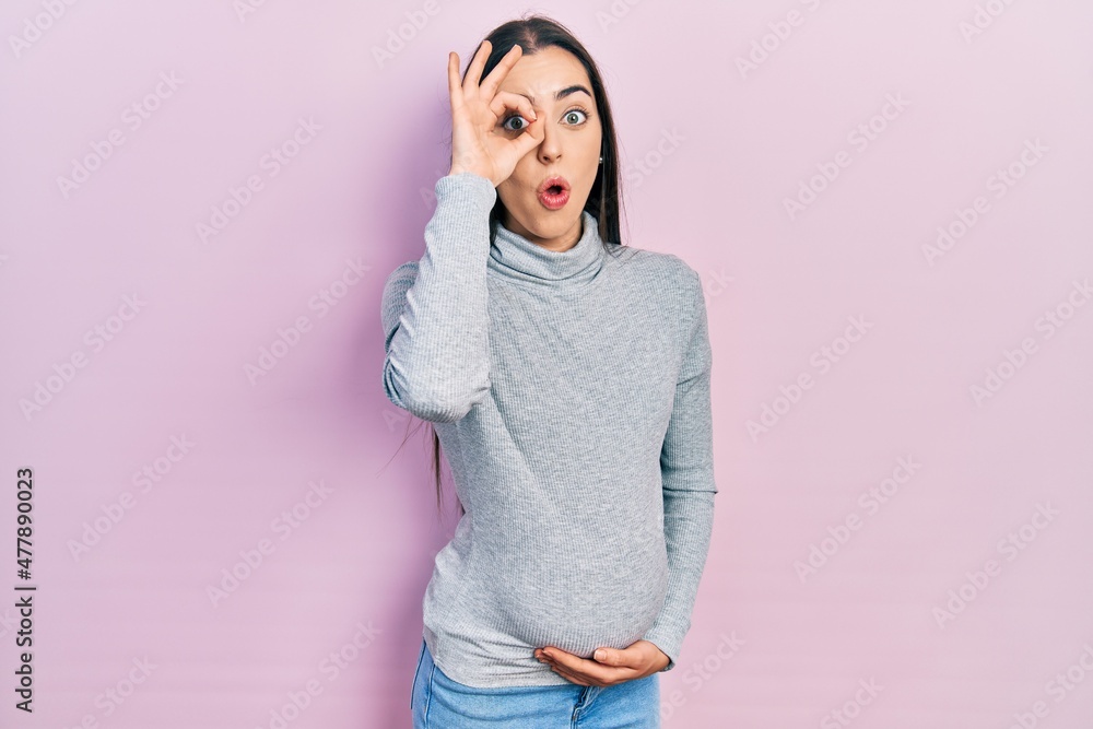 Beautiful woman with blue eyes expecting a baby, touching pregnant belly doing ok gesture shocked with surprised face, eye looking through fingers. unbelieving expression. Beautiful woman with blue eyes expecting a baby, touching pregnant belly doing ok gesture shocked with surprised face, eye looking through fingers. unbelieving expression.