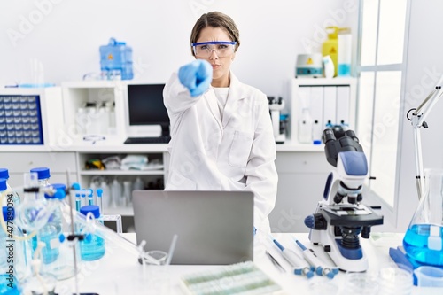 Young hispanic woman wearing scientist uniform working at laboratory pointing with finger to the camera and to you, confident gesture looking serious
