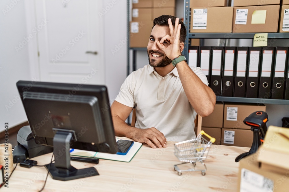 Handsome hispanic man working at small business commerce doing ok gesture with hand smiling, eye looking through fingers with happy face. Handsome hispanic man working at small business commerce doing ok gesture with hand smiling, eye looking through fingers with happy face.