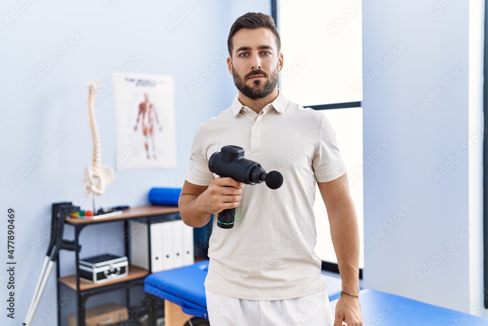 Handsome hispanic man holding therapy massage gun at physiotherapy center thinking attitude and sober expression looking self confident