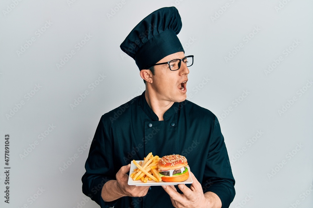 Handsome young man chef holding burger with fries angry and mad screaming frustrated and furious, shouting with anger. rage and aggressive concept. Handsome young man chef holding burger with fries angry and mad screaming frustrated and furious, shouting with anger. rage and aggressive concept.