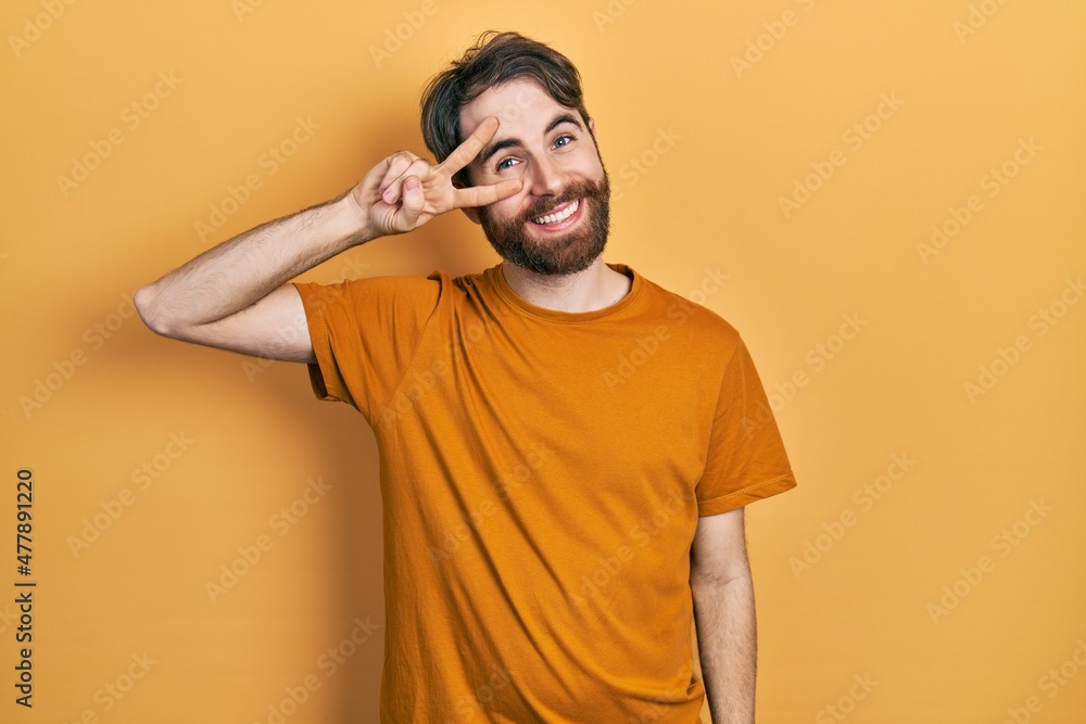 Caucasian man with beard wearing casual yellow t shirt doing peace symbol with fingers over face, smiling cheerful showing victory Caucasian man with beard wearing casual yellow t shirt doing peace symbol with fingers over face, smiling cheerful showing victory