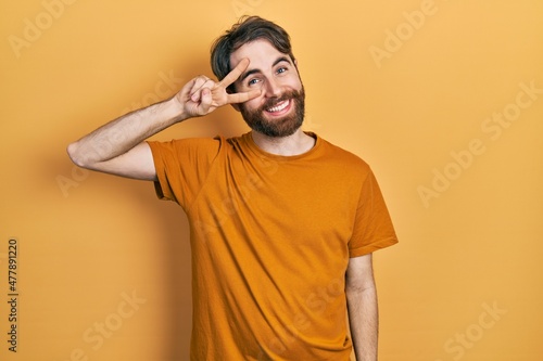 Caucasian man with beard wearing casual yellow t shirt doing peace symbol with fingers over face, smiling cheerful showing victory