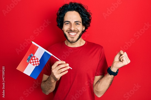 Handsome hispanic man holding croatia flag screaming proud, celebrating victory and success very excited with raised arm