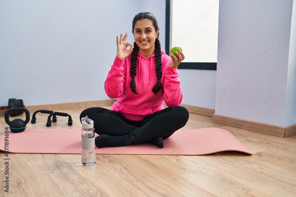 Young hispanic woman sitting on yoga mat eating apple doing ok sign with fingers, smiling friendly gesturing excellent symbol Young hispanic woman sitting on yoga mat eating apple doing ok sign with fingers, smiling friendly gesturing excellent symbol
