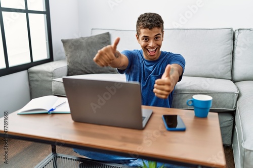 Young handsome hispanic man using laptop sitting on the floor approving doing positive gesture with hand, thumbs up smiling and happy for success. winner gesture.