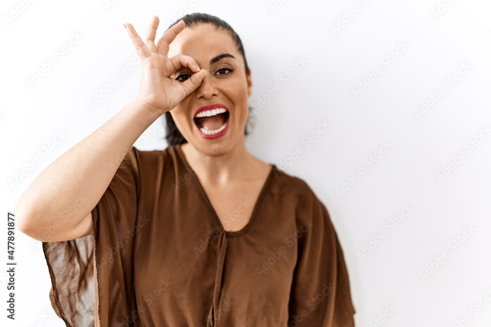 Young brunette woman standing over isolated background doing ok gesture with hand smiling, eye looking through fingers with happy face. Young brunette woman standing over isolated background doing ok gesture with hand smiling, eye looking through fingers with happy face.