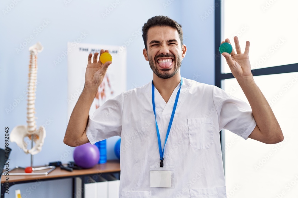 Young hispanic physiotherapist man holding strength balls to train hand muscles sticking tongue out happy with funny expression. Young hispanic physiotherapist man holding strength balls to train hand muscles sticking tongue out happy with funny expression.