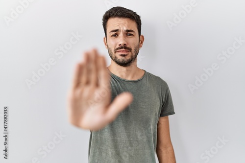 Young hispanic man with beard wearing casual t shirt over white background doing stop sing with palm of the hand. warning expression with negative and serious gesture on the face.