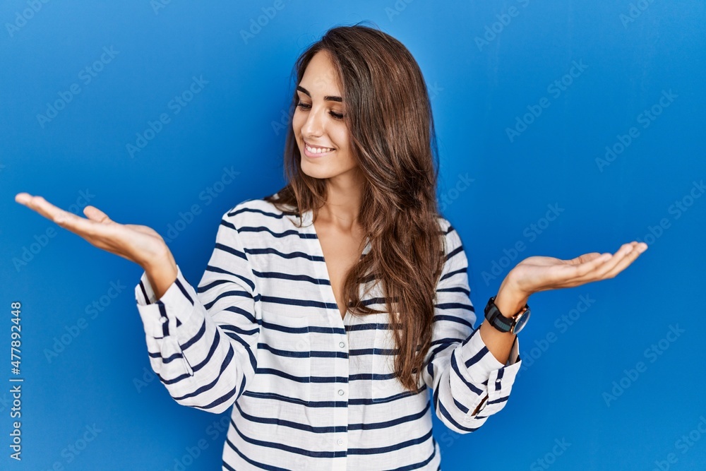 Young hispanic woman standing over blue isolated background smiling showing both hands open palms, presenting and advertising comparison and balance Young hispanic woman standing over blue isolated background smiling showing both hands open palms, presenting and advertising comparison and balance