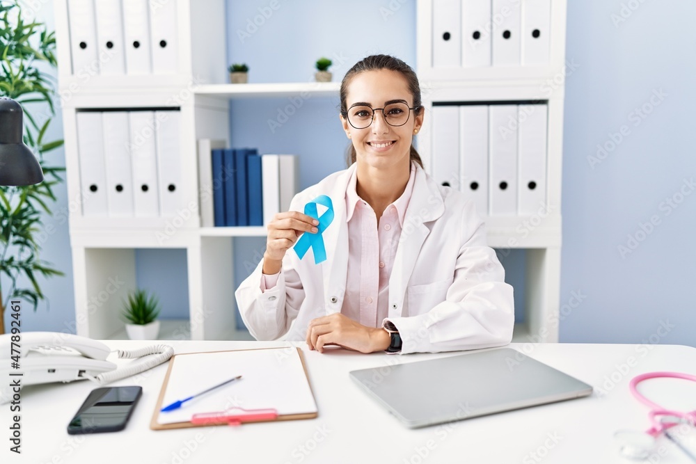 Young hispanic woman wearing doctor uniform holding blue ribbon at clinic Young hispanic woman wearing doctor uniform holding blue ribbon at clinic