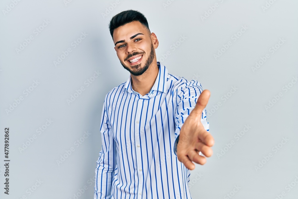 Young hispanic man with beard wearing casual striped shirt smiling friendly offering handshake as greeting and welcoming. successful business. Young hispanic man with beard wearing casual striped shirt smiling friendly offering handshake as greeting and welcoming. successful business.