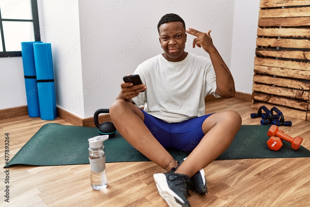 Young african man sitting on training mat at the gym using smartphone shooting and killing oneself pointing hand and fingers to head like gun, suicide gesture. Young african man sitting on training mat at the gym using smartphone shooting and killing oneself pointing hand and fingers to head like gun, suicide gesture.