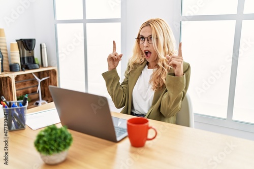 Beautiful blonde woman working at the office with laptop amazed and surprised looking up and pointing with fingers and raised arms.