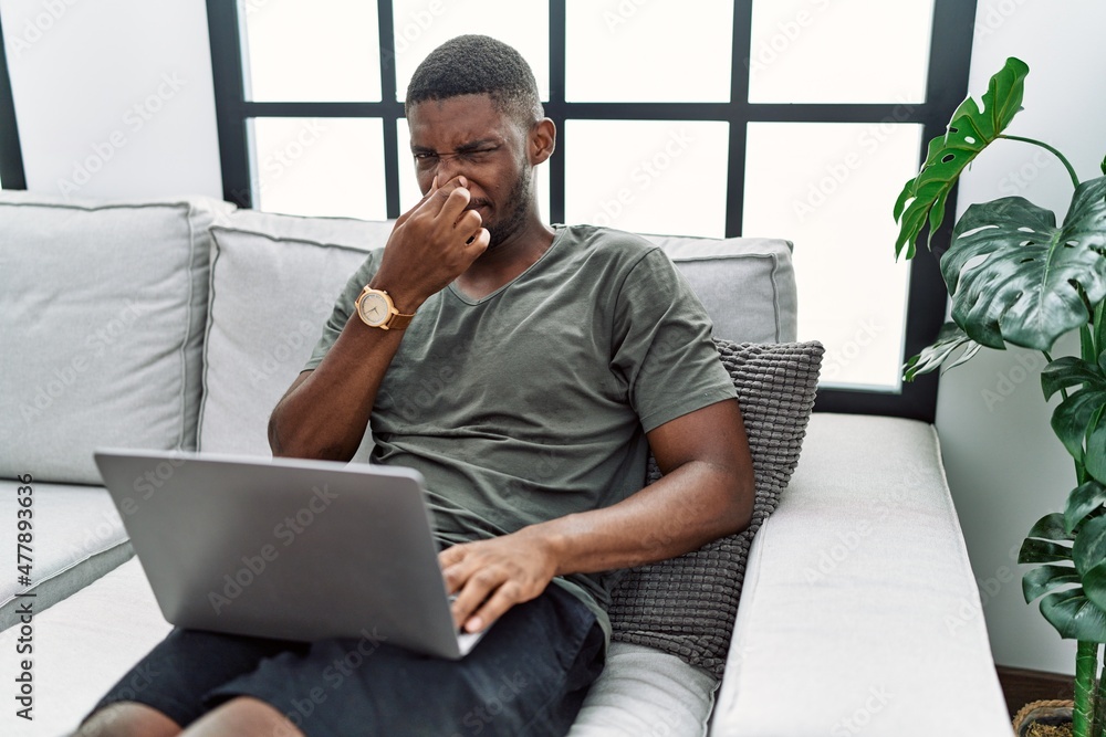 Young african american man using laptop at home sitting on the sofa smelling something stinky and disgusting, intolerable smell, holding breath with fingers on nose. bad smell - obrazy, fototapety, plakaty 