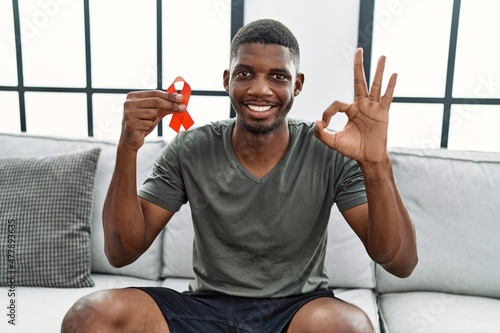 Young african american man holding support red ribbon sitting on the sofa doing ok sign with fingers  smiling friendly gesturing excellent symbol